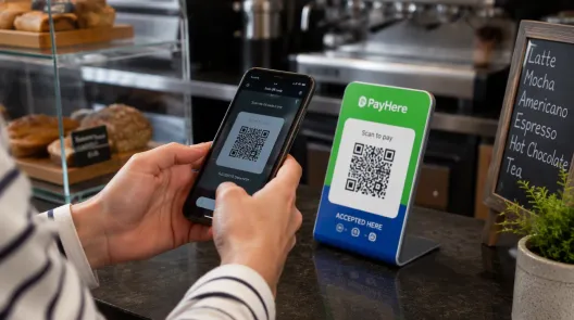 Persona escaneando un código QR de pago con su celular en el mostrador de una cafetería, junto a un cartel que dice PayHere. Scan to pay
